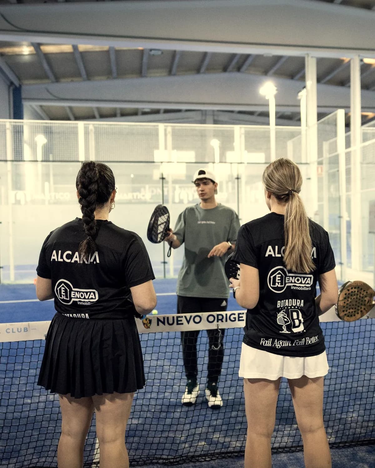 A coach explains a drill to two players, seen from behind, at the net of an indoor court.