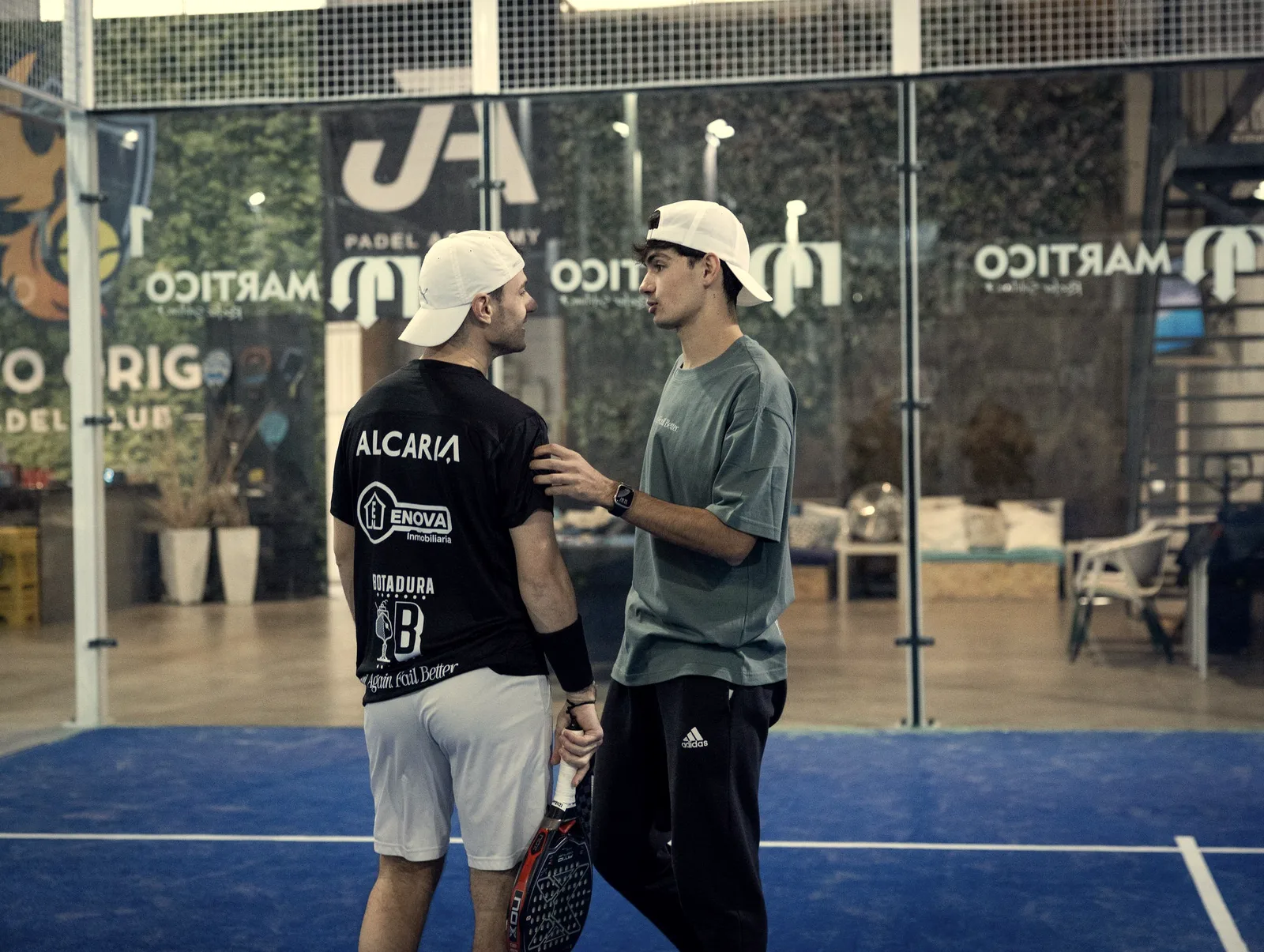 Un entrenador de gorra blanca y camiseta verde habla de cerca a un alumno de espaldas, con la mano en su hombro, en una pista cubierta