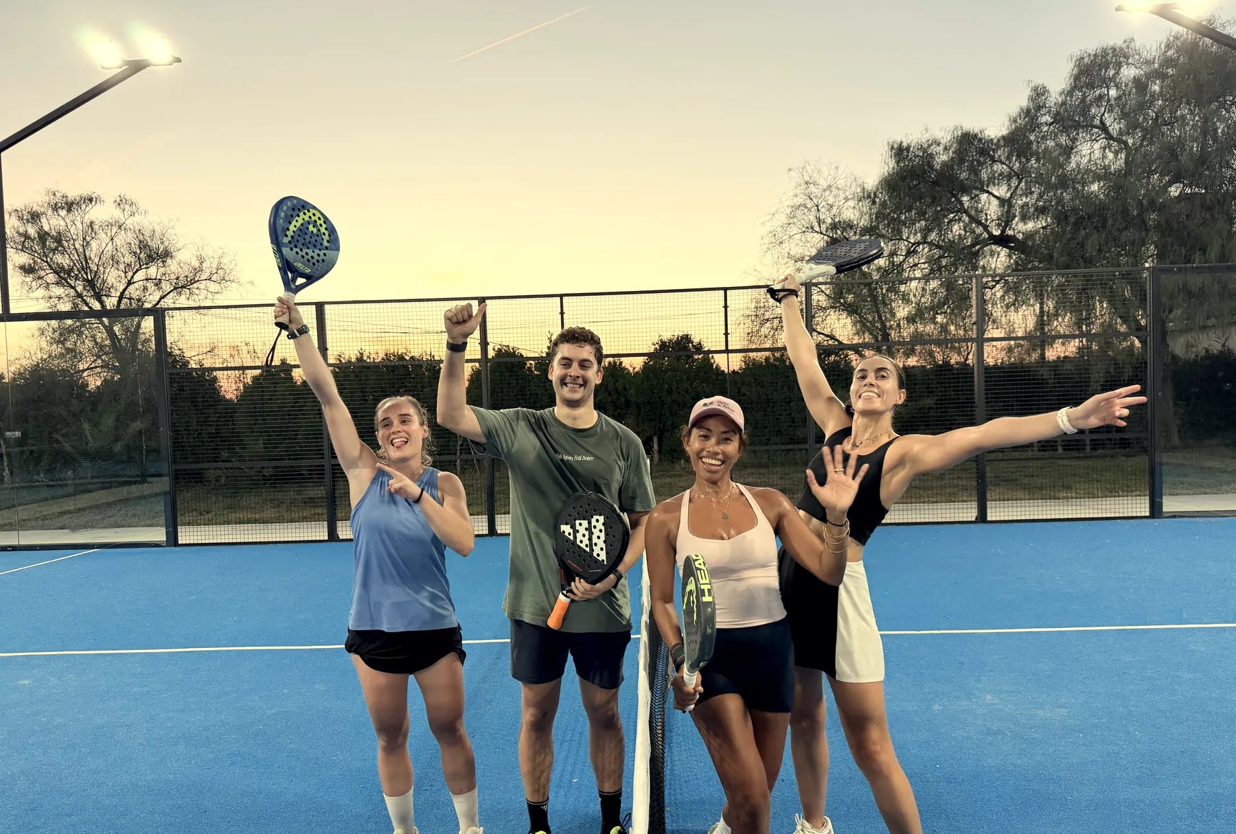 Cuatro jugadores celebran juntos junto a la red de una pista al atardecer, con los brazos en alto
