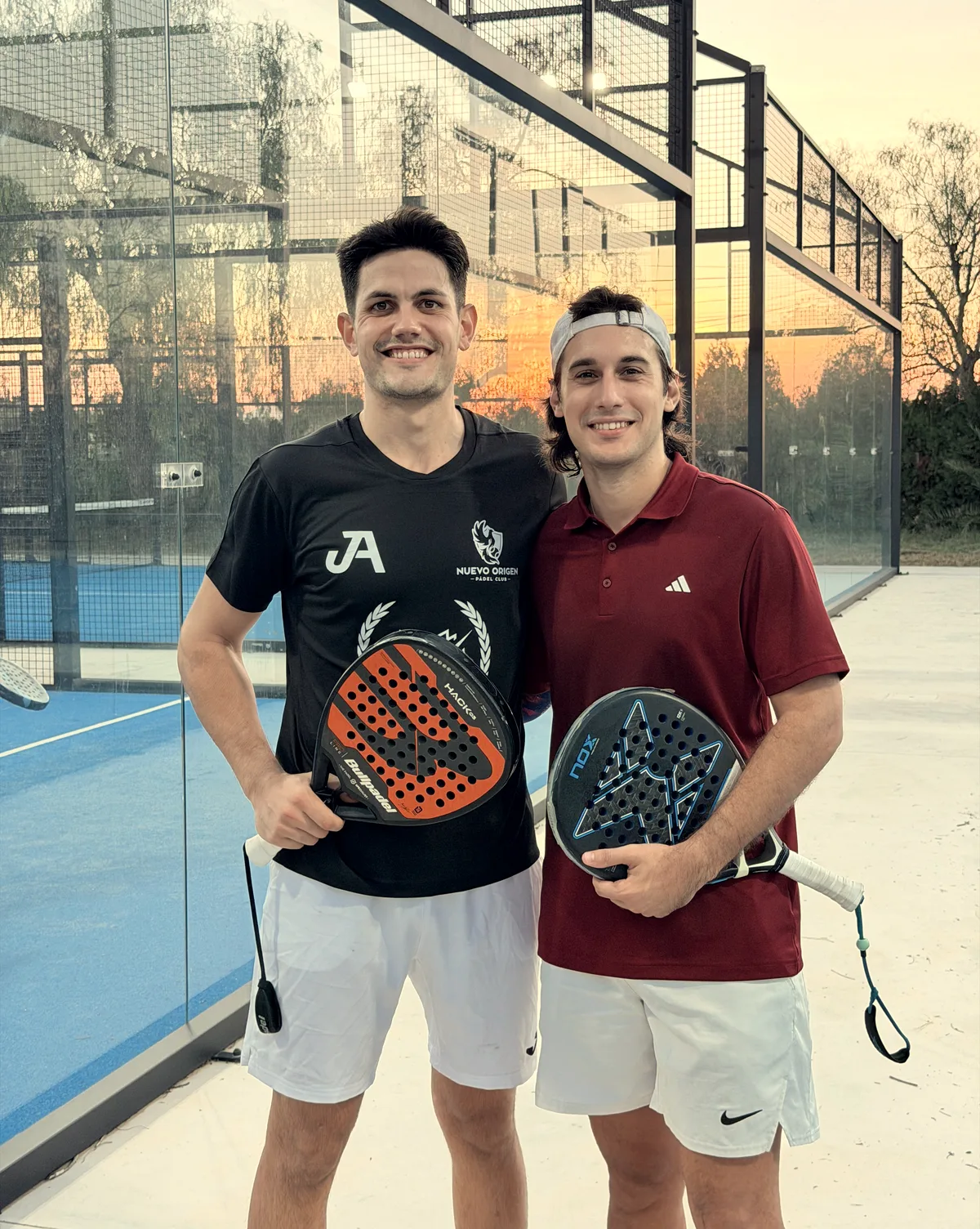Dos jugadores sonríen con las palas al atardecer: uno con la camiseta de la academia y otro con polo granate