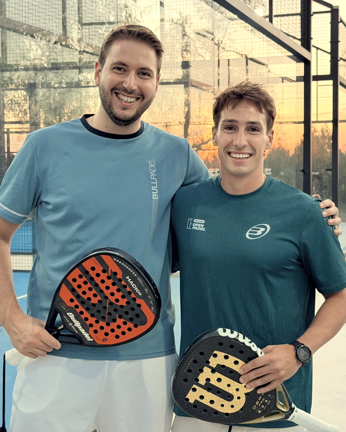 Dos jugadores sonríen abrazados en el pasillo entre pistas, con el atardecer detrás