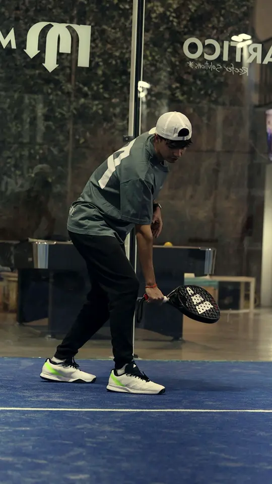 Un entrenador de JA con gorra blanca baja la pala hacia una bola baja en una pista azul cubierta.
