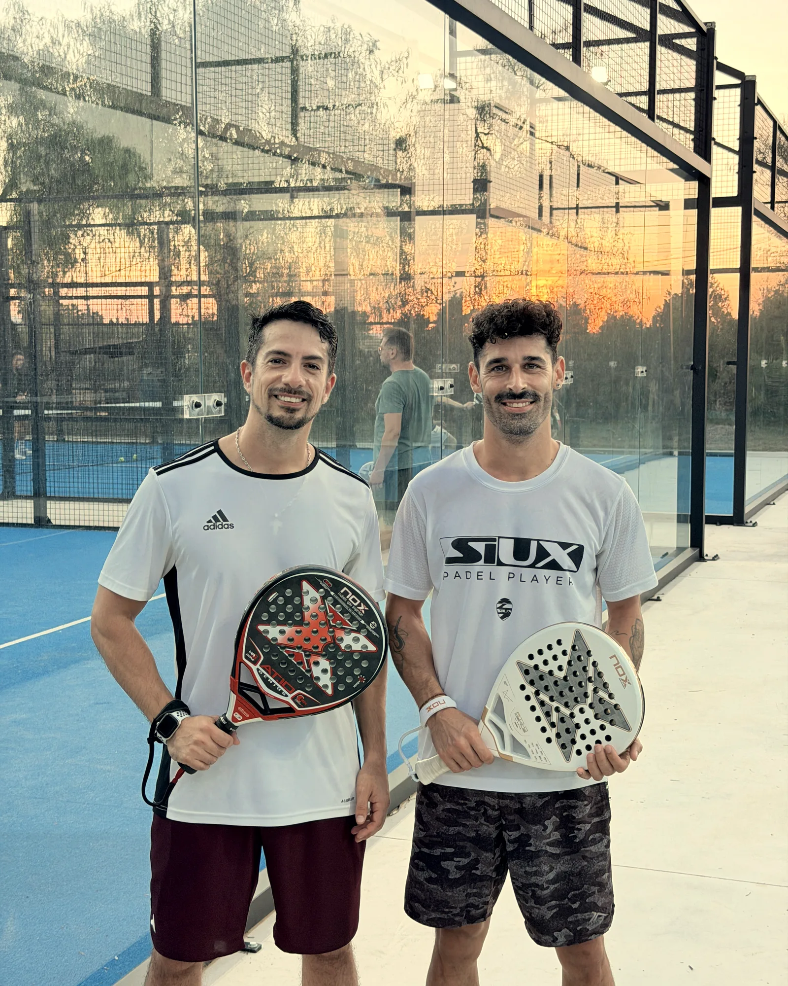 Two players in white T-shirts smile, rackets in hand, on a glass court at sunset