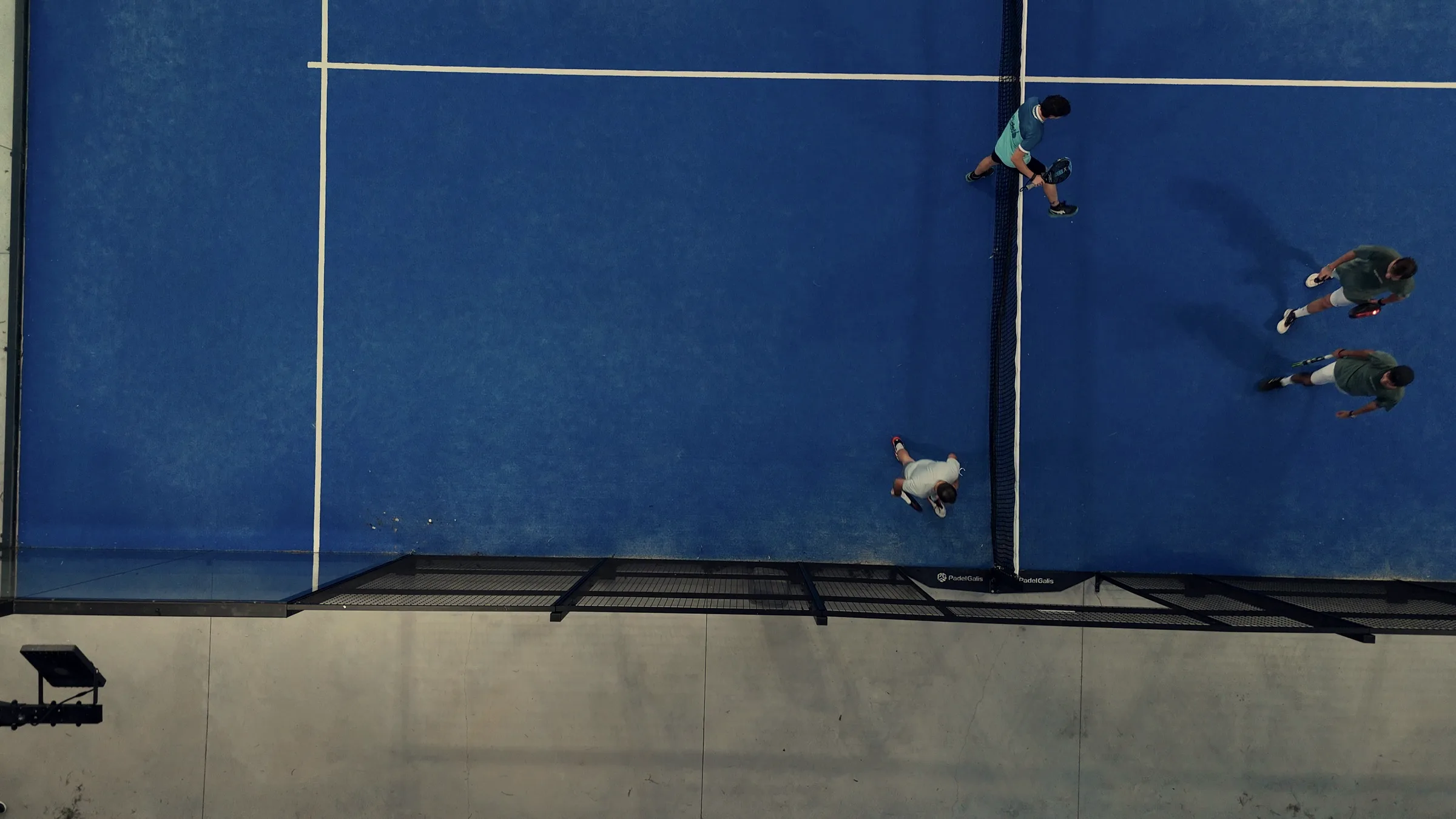 A padel court seen from above, with the net, the glass and three players