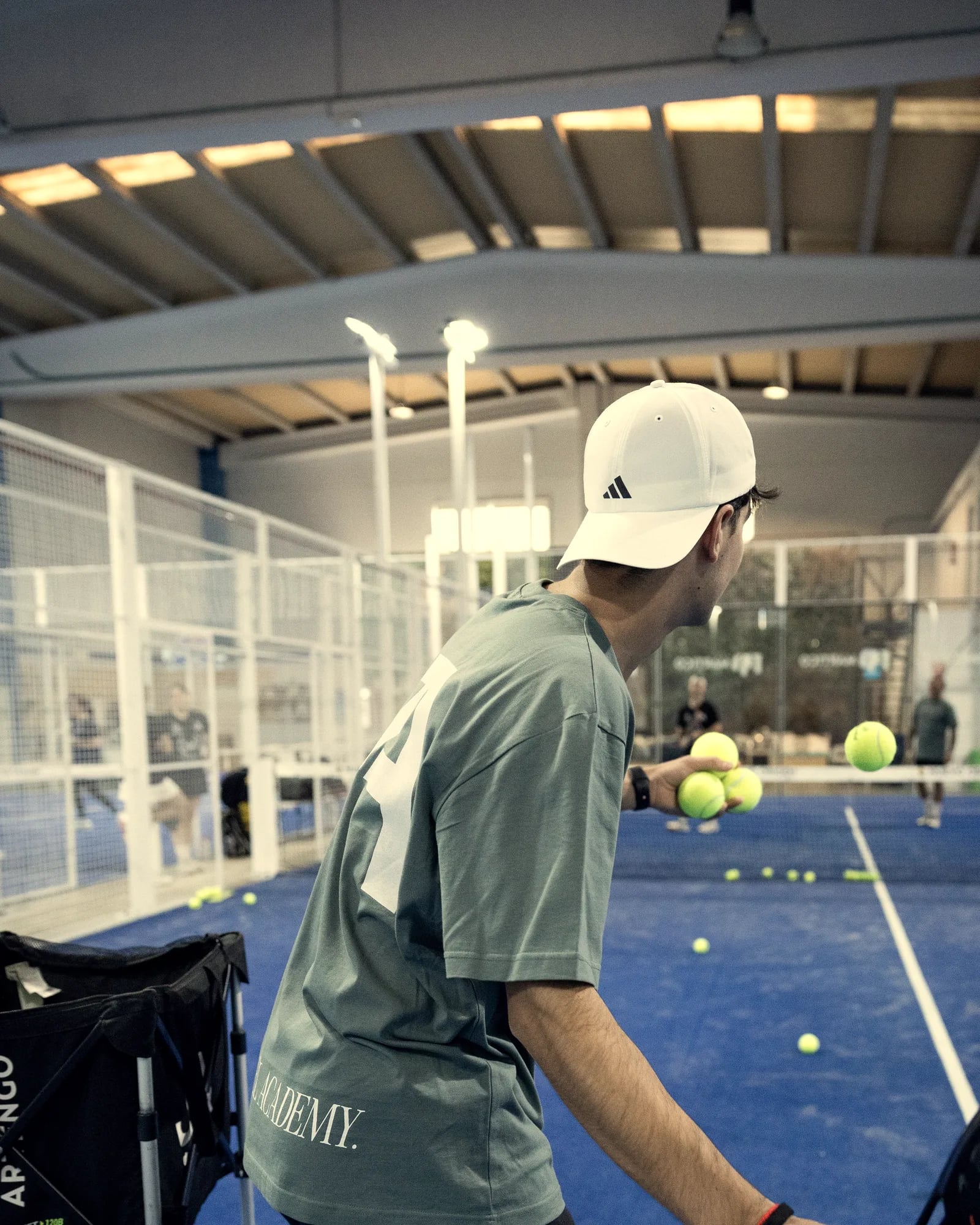 Un entrenador da bola desde el carro en una pista azul llena de bolas, bajo el techo de madera de la nave de Origen Padel Club